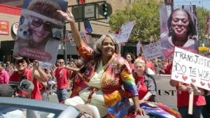 Miss Major Griffin-Gracy is sitting in a convertible and waving. She is surrounded by people at a Pride parade.